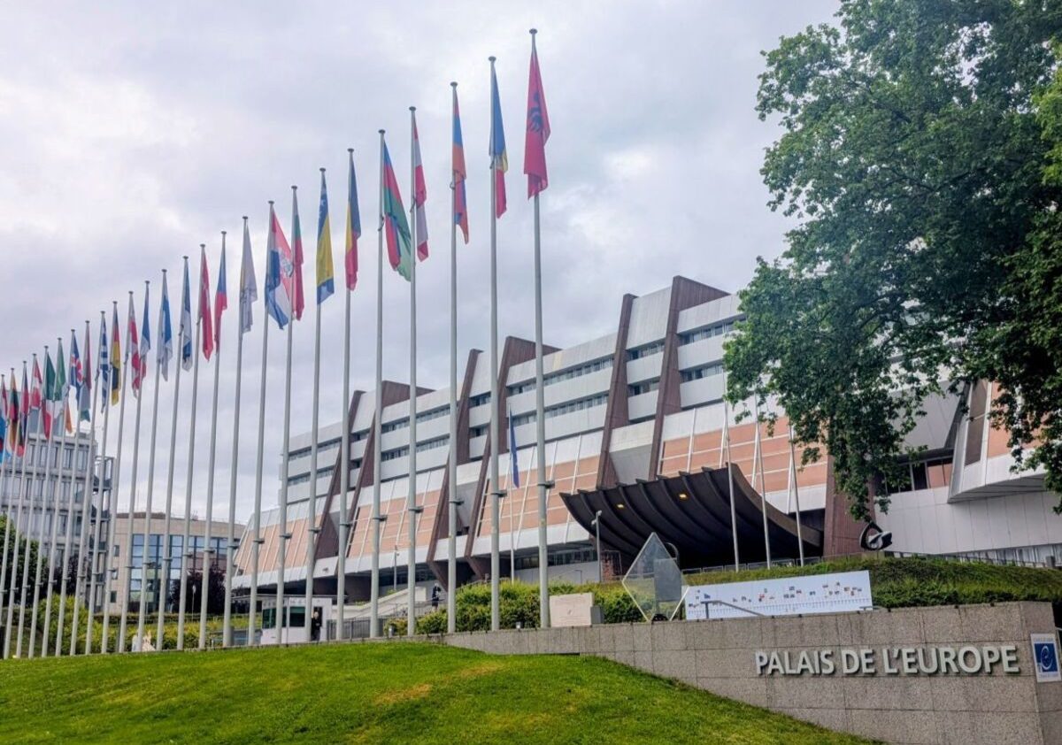 Exterior view of the Palais de l'Europe in Strasbourg, with a long row of national flagpoles and a stone sign in the foreground