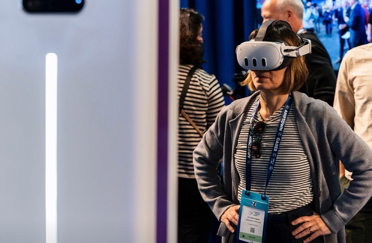 A visitor wearing a Meta Quest VR headset stands at an immersive technology trade show demo in front of a volumetric display booth