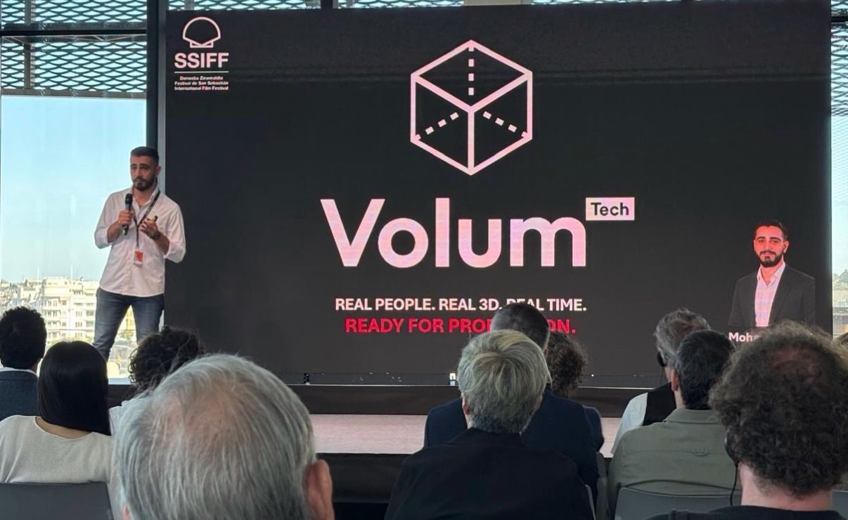 A speaker presenting on stage at the San Sebastián Film Festival, in front of a large screen showing the Volum Tech logo and the tagline “Real people. Real 3D. Real time.”